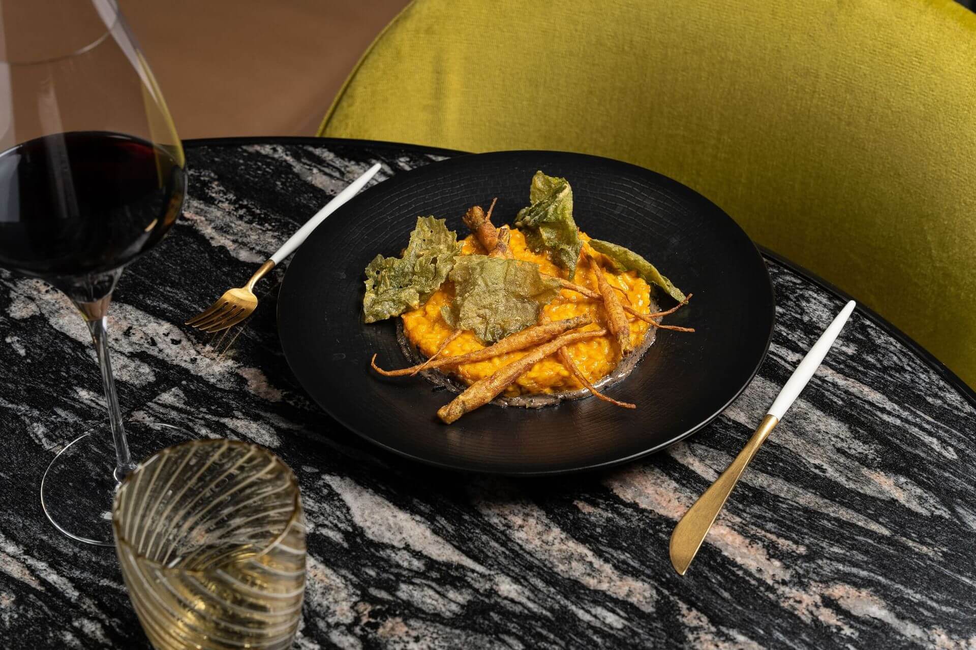 A black plate of gourmet risotto topped with crispy root vegetables and greens, served on a marble table with a glass of red wine and a glass of water.