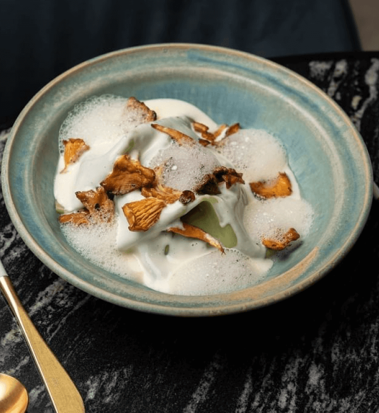 A blue ceramic bowl from Virens Restaurant holds a creamy white sauce, delicate foam, and golden-brown mushrooms, elegantly placed on a black marble surface with a gold spoon beside it.
