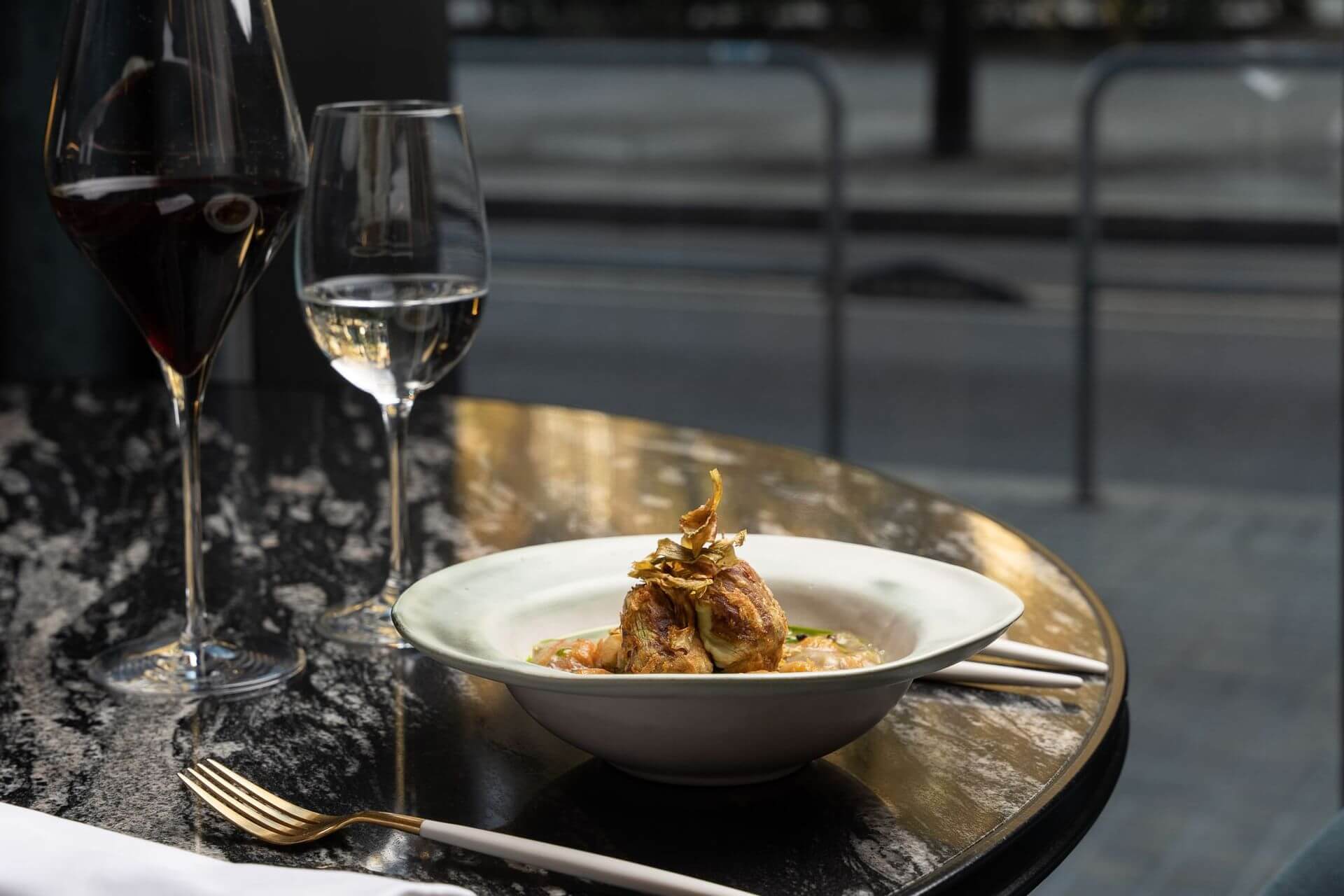 A white plate with a gourmet dish, accompanied by glasses of red and white wine, on a marble table by a window overlooking a street.