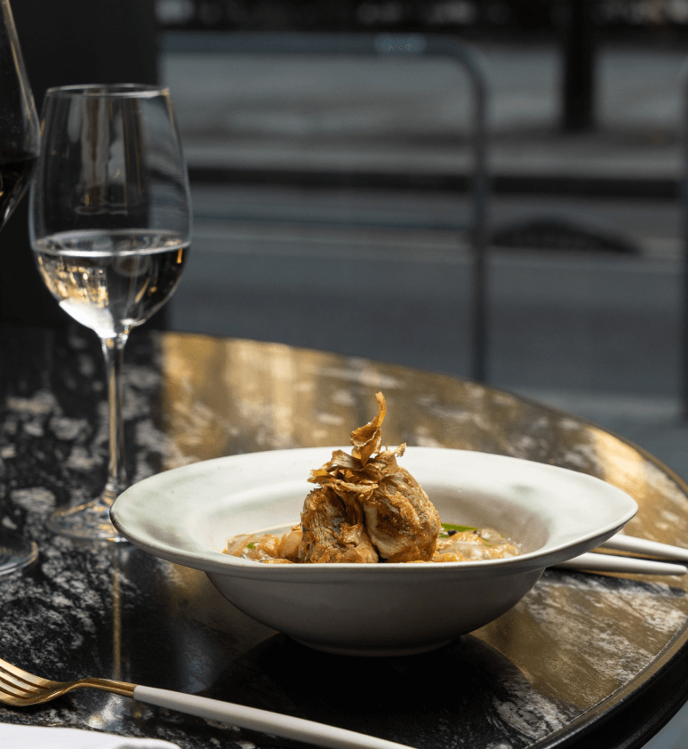 A plated gourmet dish with garnished meat and sauce sits on a marble table at Virens Restaurant, accompanied by two glasses of wine in a stylish restaurant setting by a window.