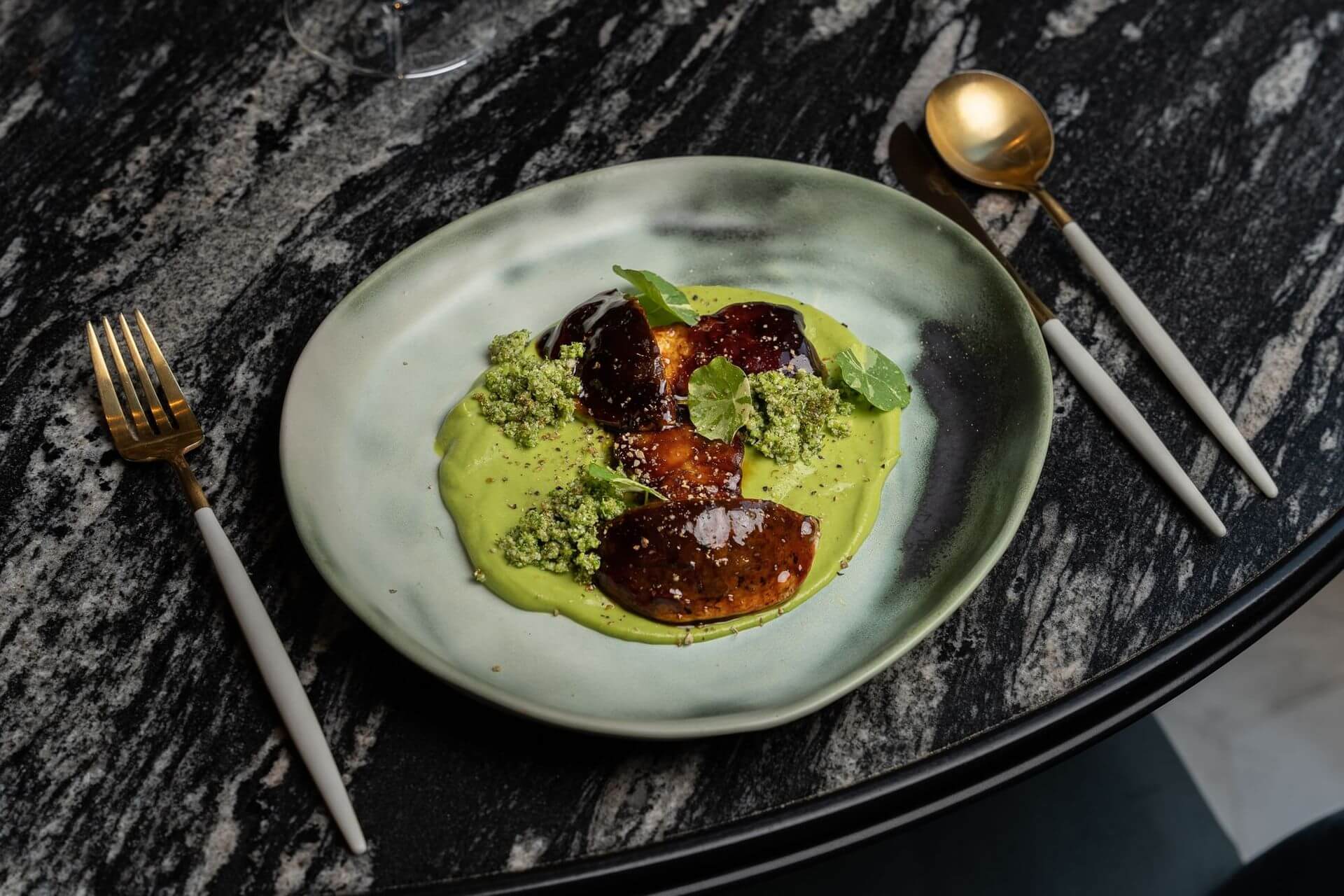 A ceramic plate with three glazed pieces of food on a green puree, garnished with green crumbs and herbs, on a marble table with gold and white cutlery.