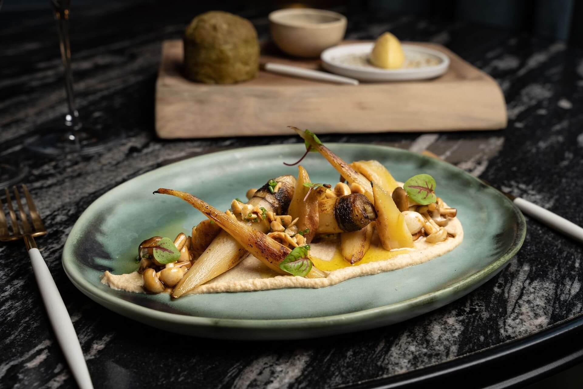 A plated dish featuring roasted carrots and mushrooms on a bed of purรฉe, garnished with nuts and microgreens, with additional food items blurred in the background.