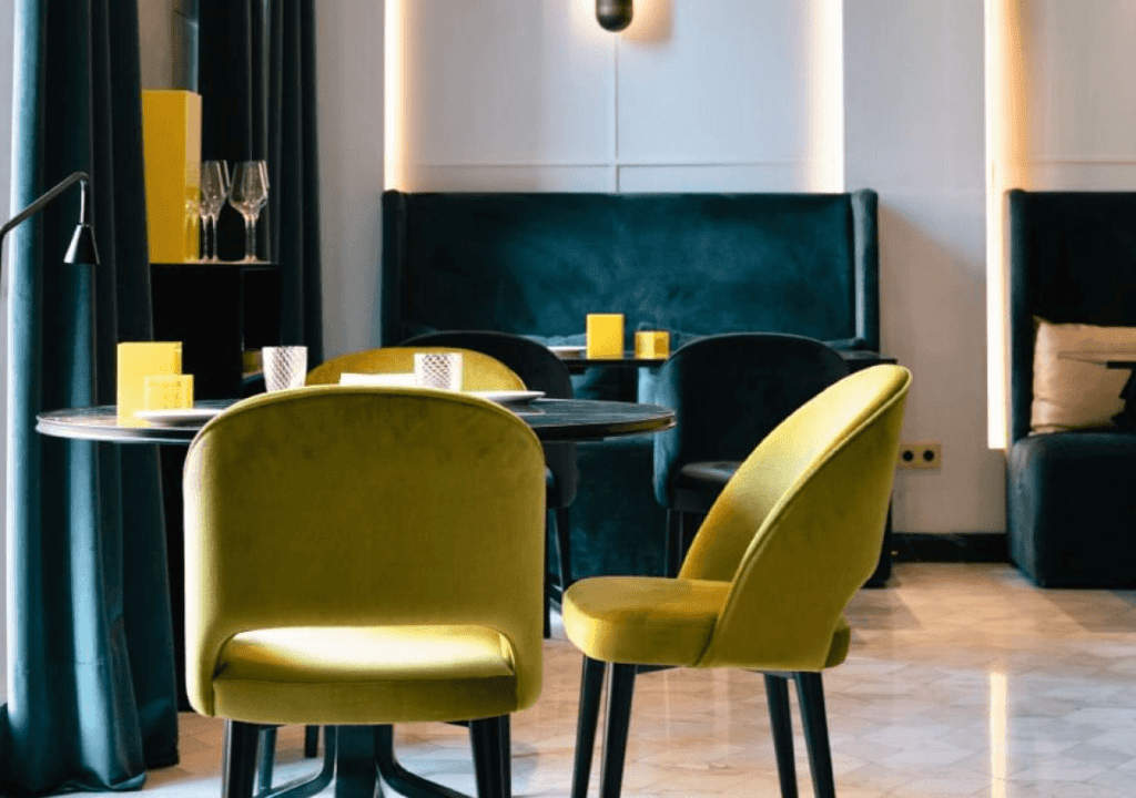 Modern Virens Restaurant interior with green velvet chairs, dark round tables, built-in dark blue seating, a marble floor, and yellow accent decor.