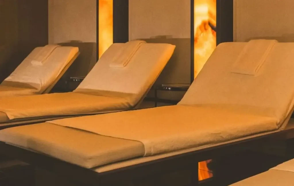 Tres tumbonas acolchadas cubiertas con toallas de color beige están dispuestas en una habitación tenuemente iluminada con cálidos paneles murales brillantes al fondo.