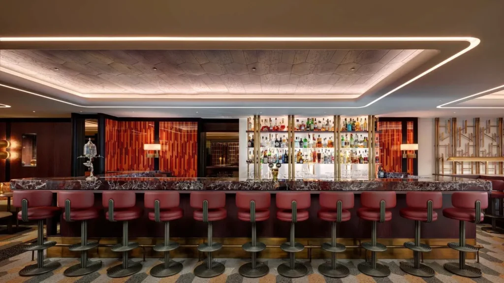 A modern bar with a marble countertop, red bar stools, and a backlit shelf displaying various drinks. Warm lighting, geometric patterns, and a curated selection of food create a welcoming atmosphere.