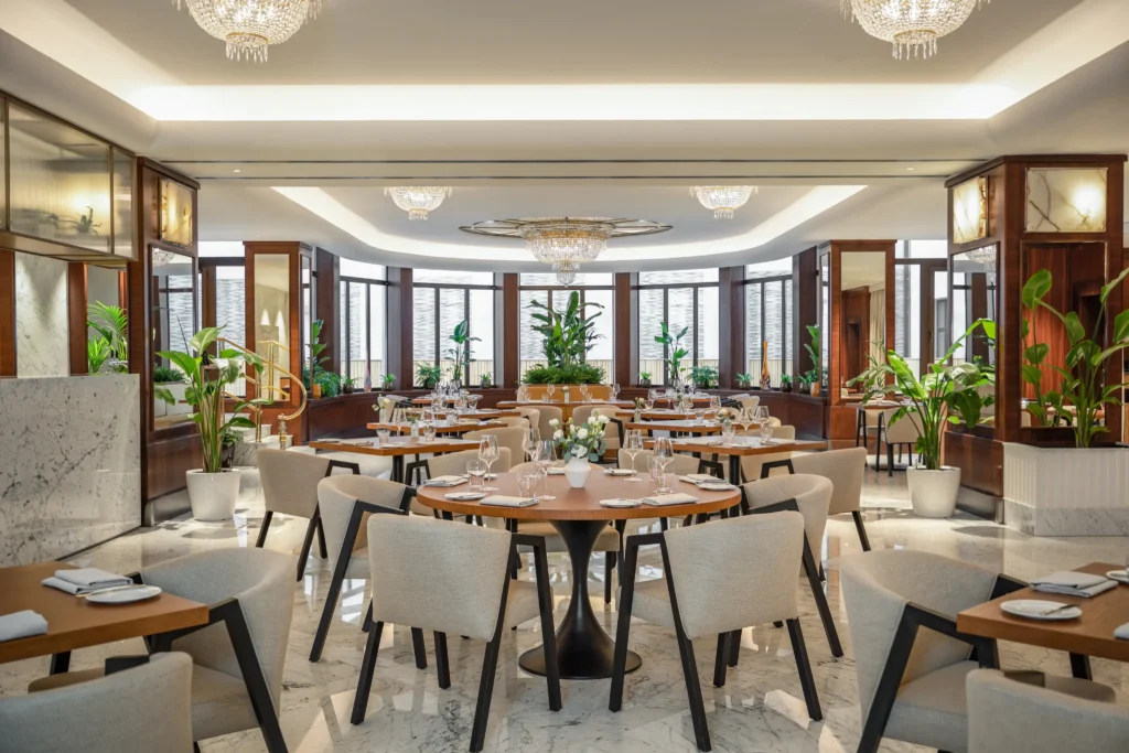 A modern, elegant restaurant dining area with round wooden tables, cream upholstered chairs, marble floors, potted plants, and large windows letting in natural light—perfect for enjoying delicious food and drinks.