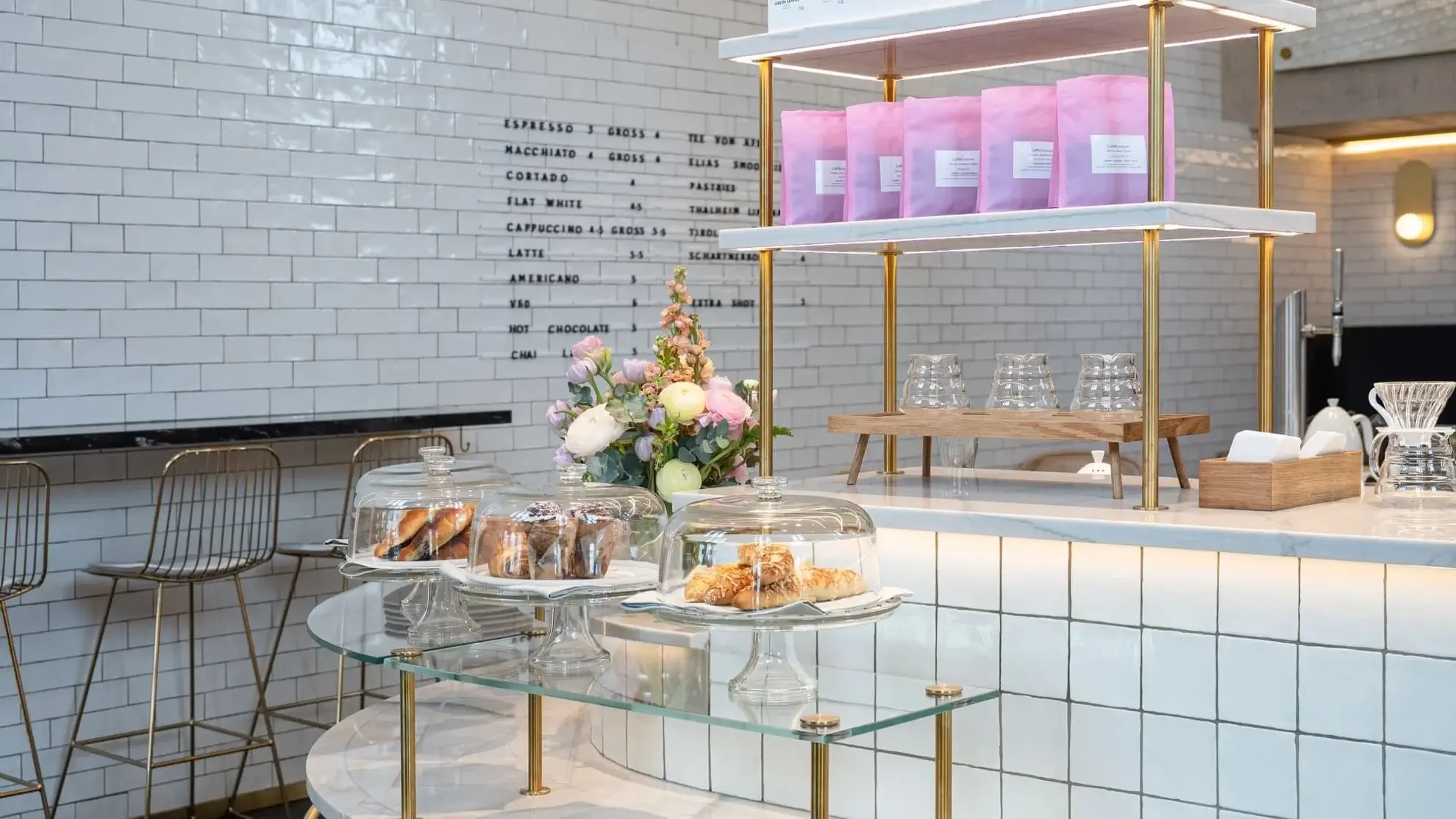 Moderní kavárenský pult s pečivem pod skleněnými kopulemi, květinovou výzdobou a růžovými sáčky s kávou na polici, na pozadí bílé kachličkové stěny s jídelním lístkem.