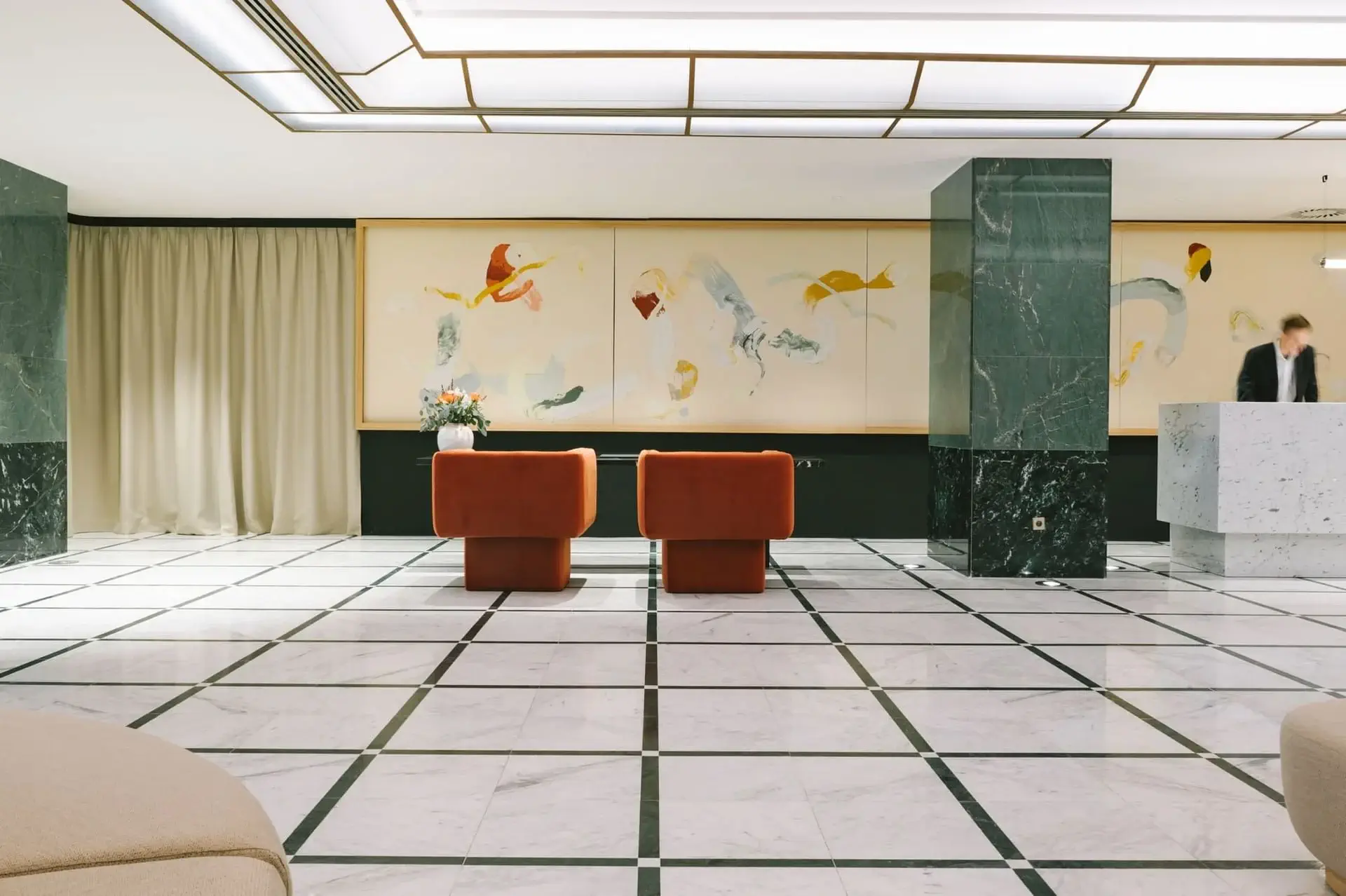 Modern hotel lobby with marble floors, two orange chairs, abstract art on the wall, and a man standing behind a white reception desk.