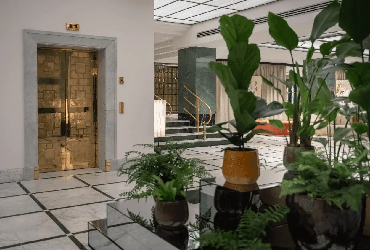 A modern lobby with marble floors, potted plants in the foreground, a gold elevator on the left, and stairs with a gold handrail in the background.