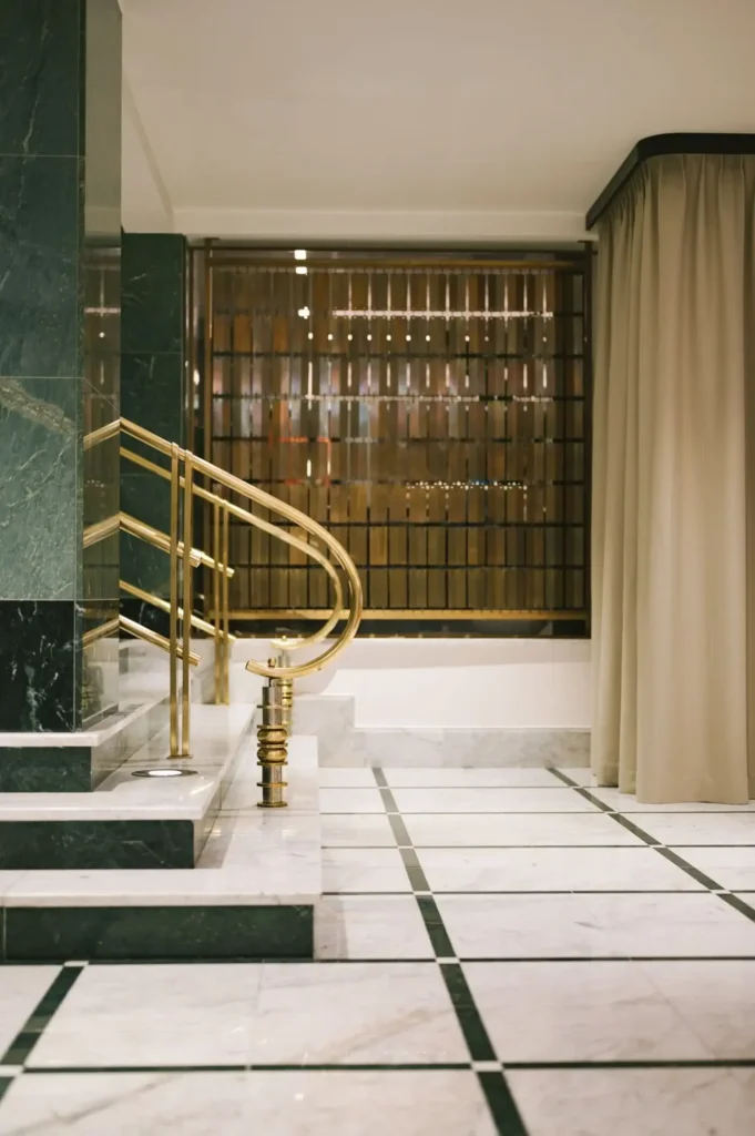A marble-tiled bathroom with gold railings, a green marble wall, beige curtains, and a window with vertical blinds above a bathtub.