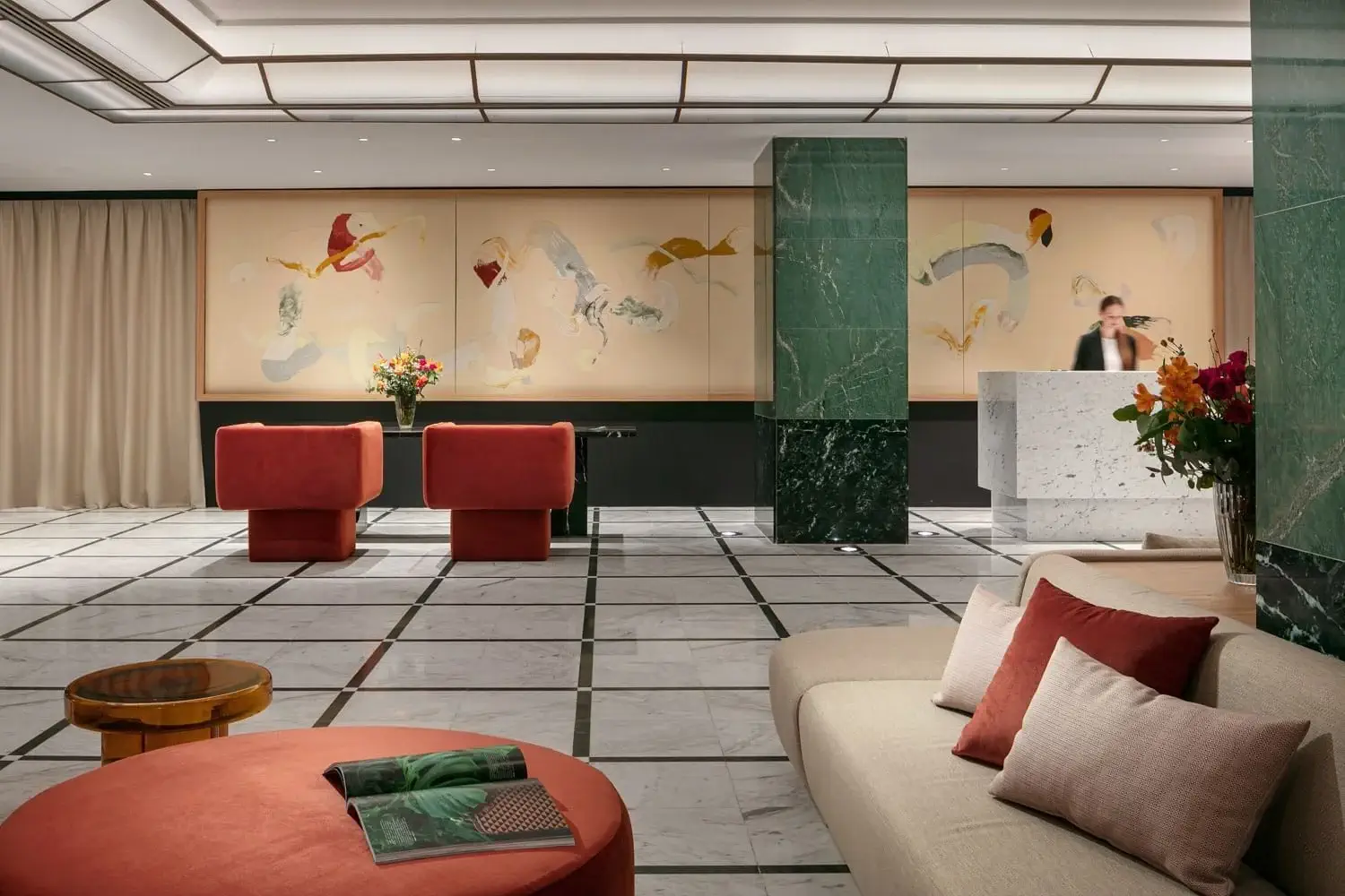 Modern hotel lobby with marble floors, green marble columns, abstract art on the wall, red seating, beige sofa, and a person at the reception desk.