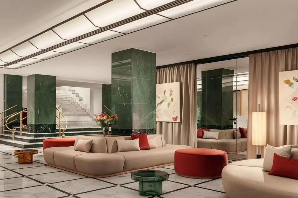 Modern hotel lobby with beige sofas, red ottomans, green marble columns, a floral arrangement, abstract artwork, and a staircase with gold railings in the background.