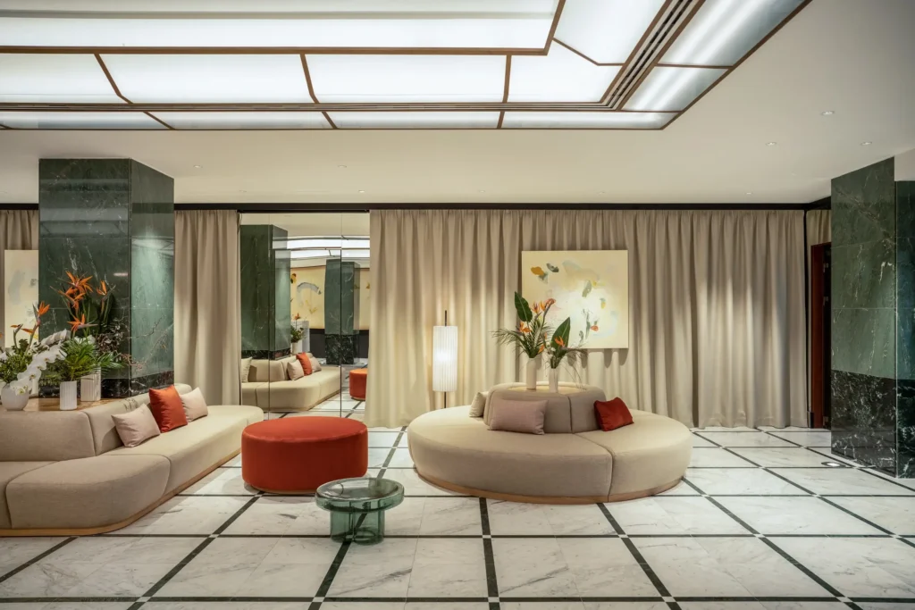 Modern hotel lobby with marble floors, green marble columns, cream sofas, red ottomans, a glass table, potted plants, neutral curtains, and abstract artwork reflecting the lively culture and happenings of the city.