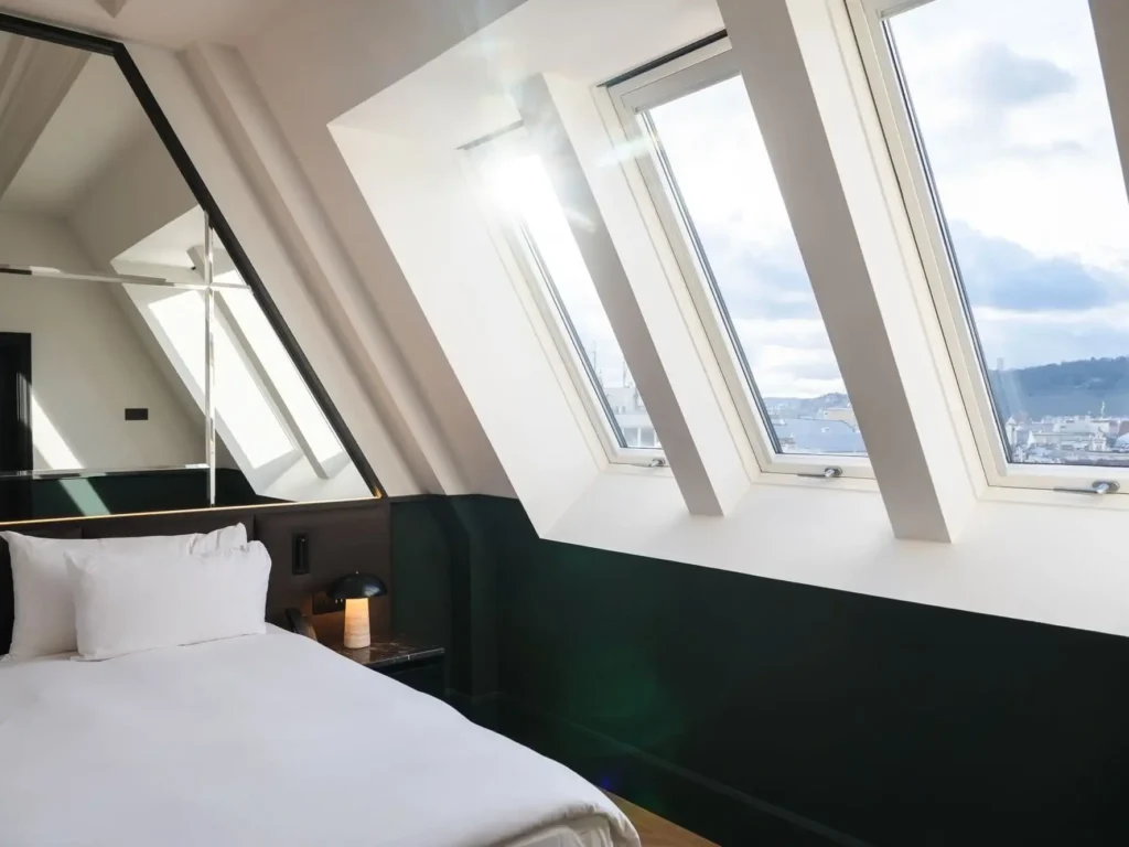 A neatly made bed beside large slanted windows with sunlight streaming in, overlooking a cityscape, in the modern Castle View Suite at Almanac X.