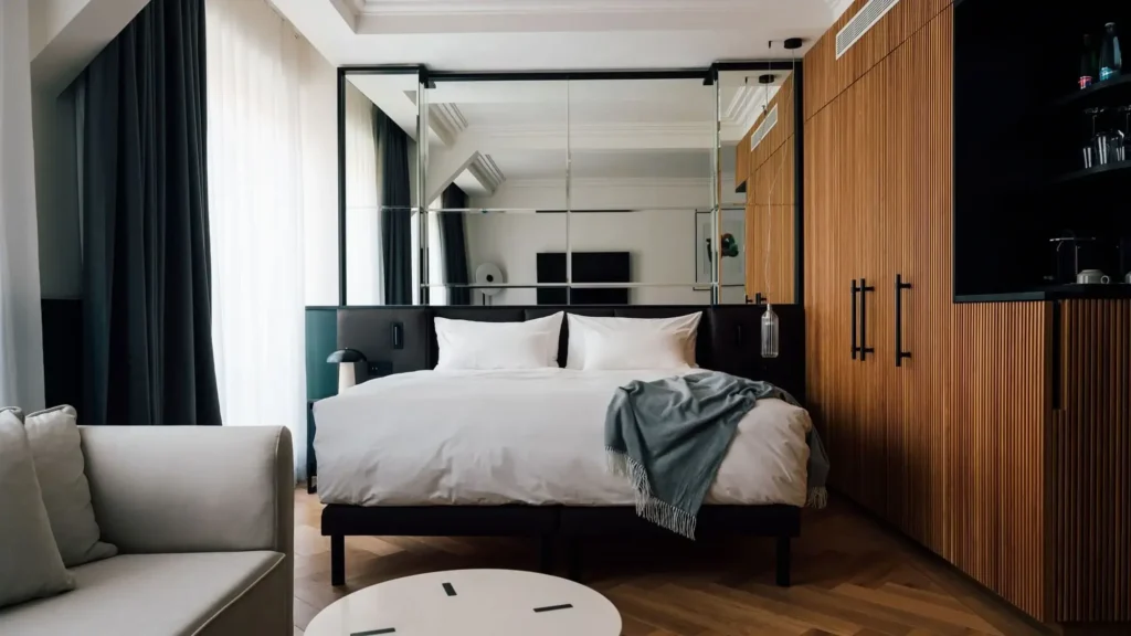Moderní Apartmá Junior má manželskou postel s bílým povlečením, šedou přehoz, velkou zrcadlovou stěnu, dřevěnou skříň a malý kulatý stolek vedle pohovky.