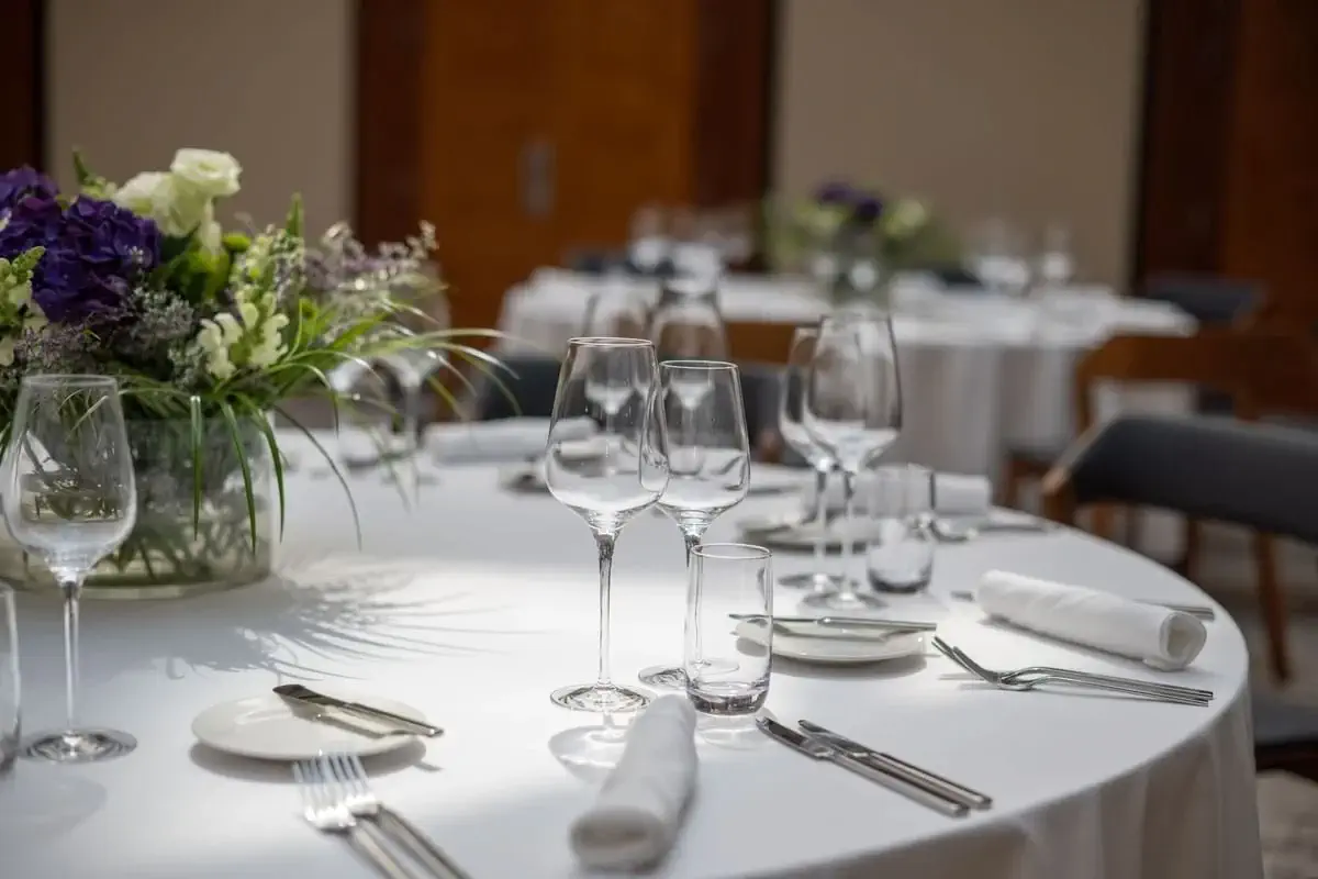 A round dining table set for a formal event with white tablecloth, glassware, plates, cutlery, and a floral centerpiece. Other similarly set tables are visible in the background.