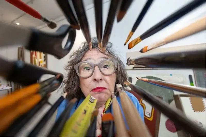 A woman with glasses looks through a circular arrangement of pencils and paintbrushes in an art studio.