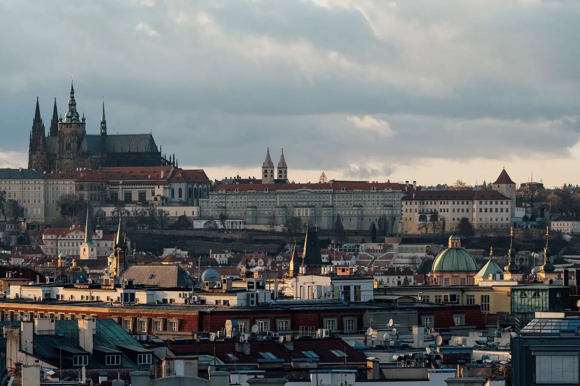 Panoramatický pohled na Prahu s Pražským hradem a katedrálou svatého Víta, s historickými budovami, střechami a módním concept store v popředí pod zataženou oblohou.
