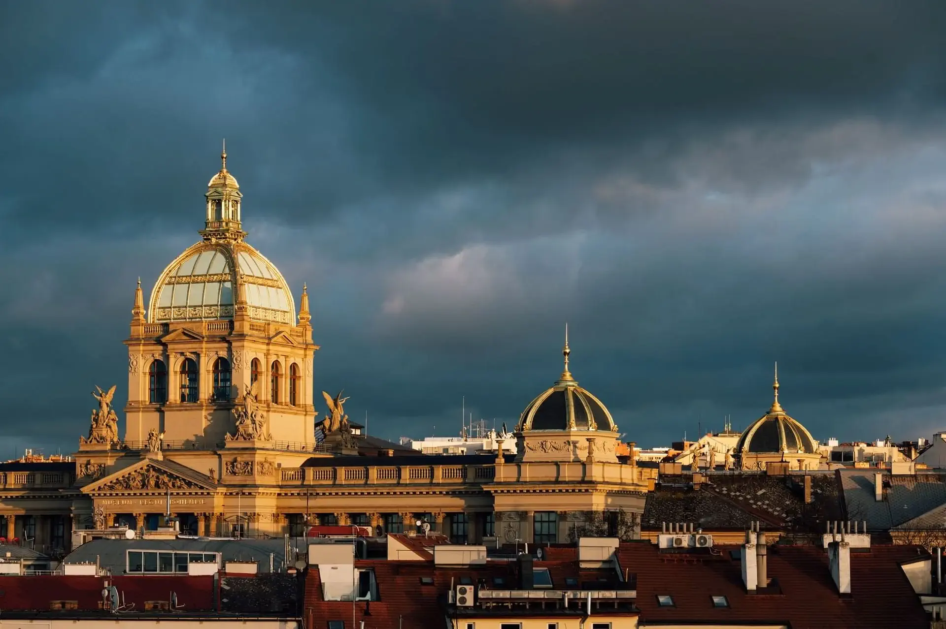 A grand historic building with domes and statues is lit by sunlight under a dramatic, cloudy sky, with red-roofed buildings and convenient parking in the foreground.