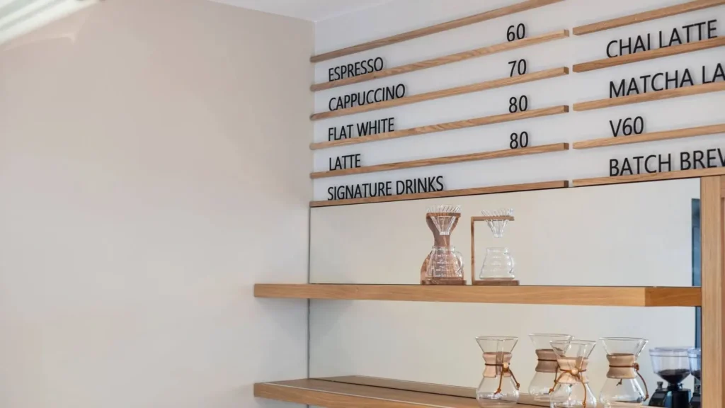 Minimalist café interior with wooden shelves holding glass coffee makers, and a menu board listing espresso, cappuccino, flat white, latte, specialty drinks, and food options with their prices.