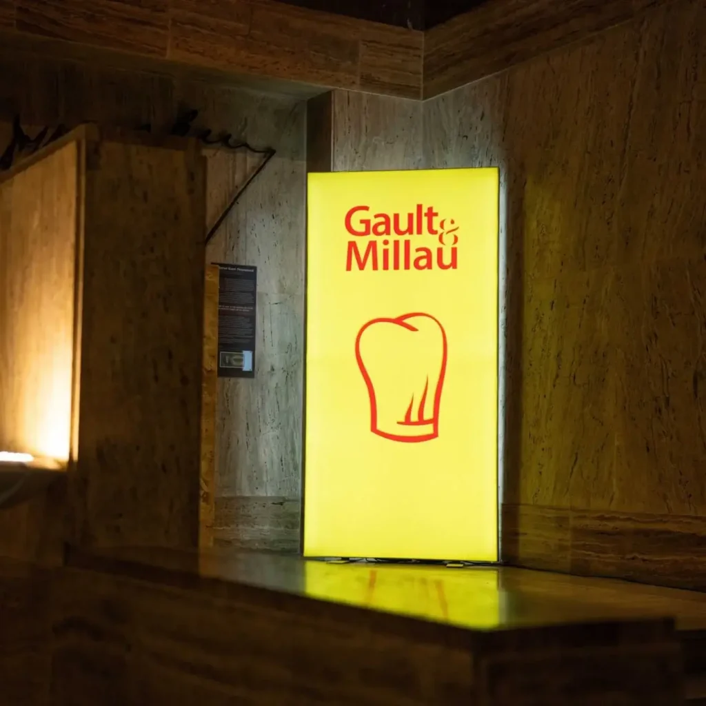 Eine gelbe Leuchtreklame mit dem roten Logo des Gault&Millau 2025, der Kochmütze und den Zwei Toques steht an einer Marmorwand im schwach beleuchteten Innenraum des Restaurants Alcron.