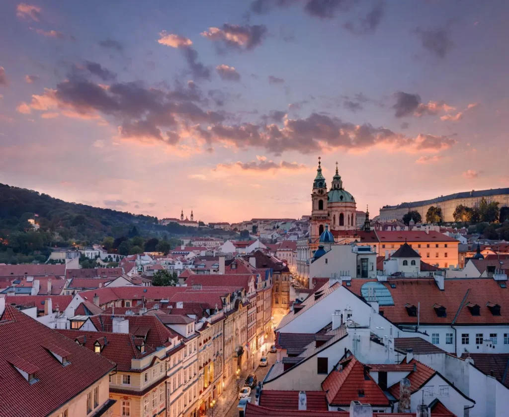 View of a European city with red-tiled rooftops, historic buildings, and church domes at sunset under a partly cloudy sky—an atmosphere perfect for Inspired Spaces.