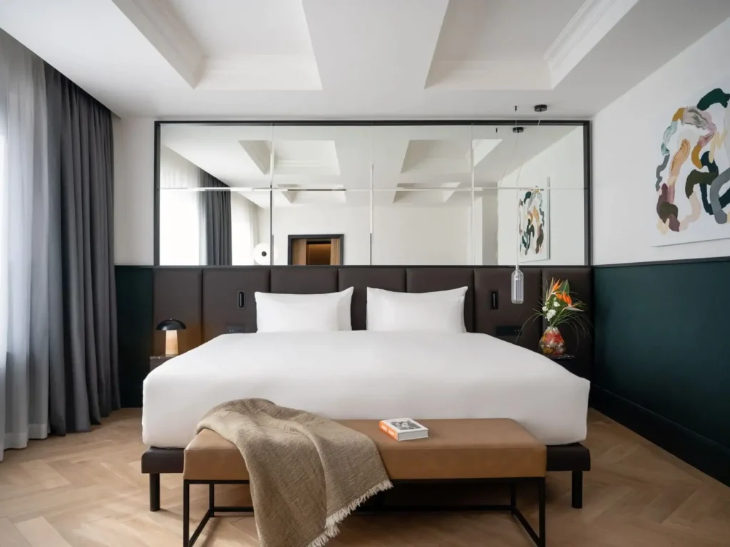 Moderní hotelový pokoj ve stylu prezidentského apartmá s velkou postelí, zrcadlovou stěnou, abstraktním uměním, tmavými závěsy, lavicí s knihou a dekou a malým stolkem s lampou a vázou s květinami.