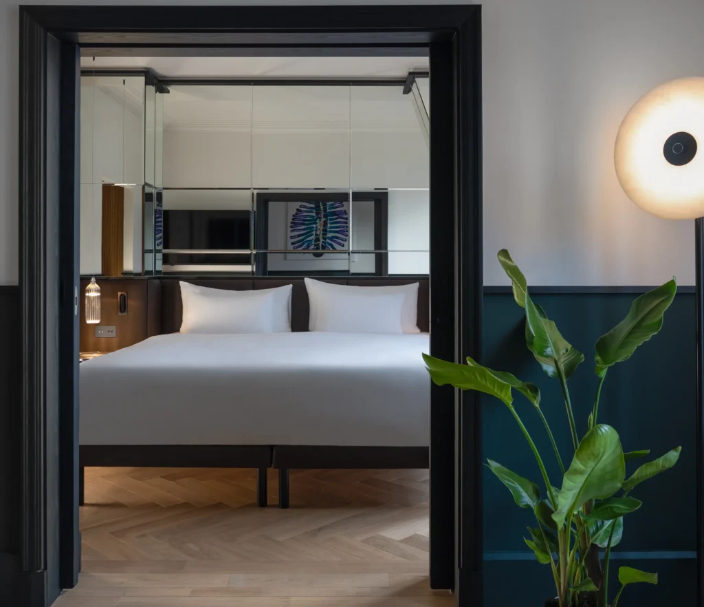 A modern Executive Suite featuring a double bed with white bedding, mirrored wall, indoor plant, round floor lamp, and elegant wood herringbone floor.