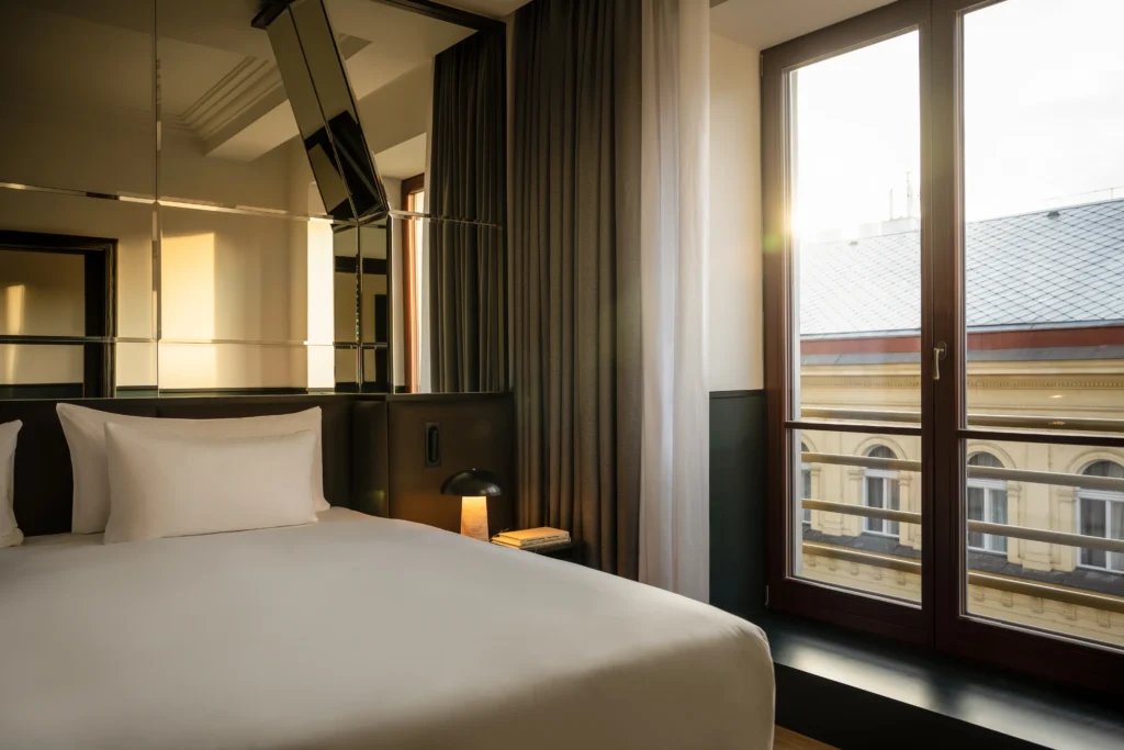 Moderní pokoj hotelu Apartmá Executive s úhledně ustlanou postelí, zrcadlovou stěnou, noční lampičkou a velkým oknem s výhledem na městskou zástavbu při západu slunce.