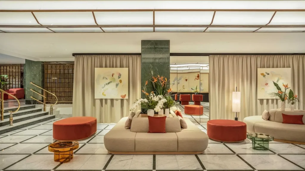 Modern hotel lobby with marble floors, neutral seating, red ottomans, floral arrangements, gold accents, and abstract art on cream-colored walls—an inviting space overseen by a dedicated Assistant Front Office Manager.