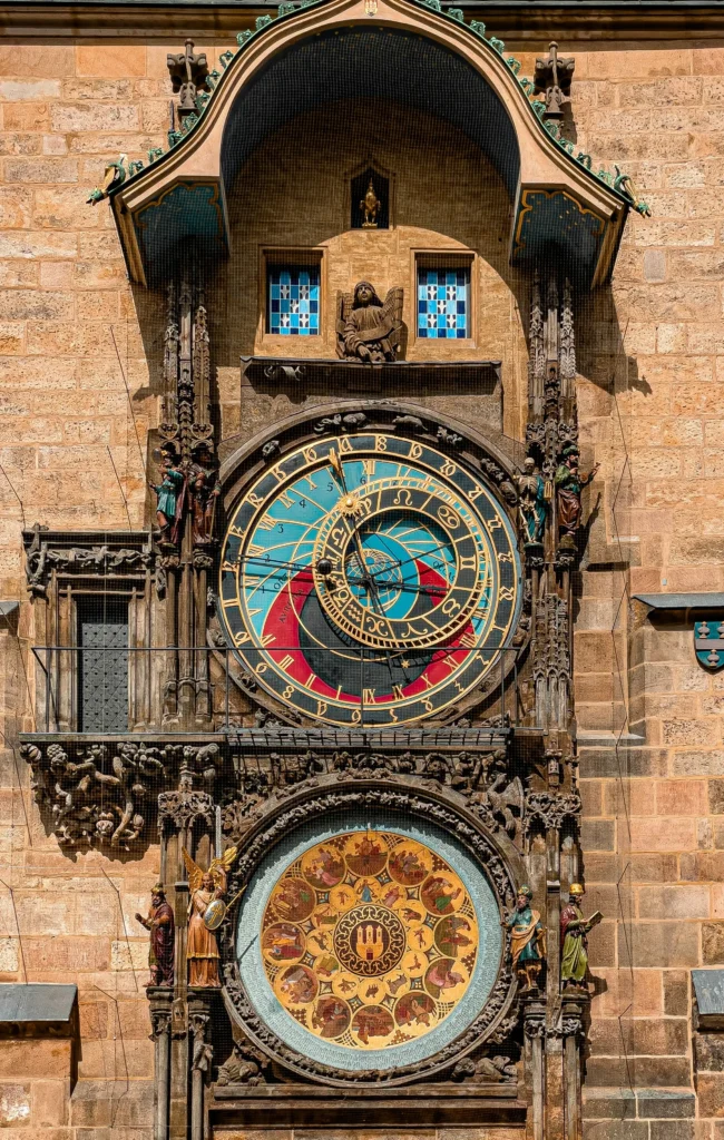 Pražský orloj na kamenné zdi s ozdobnými detaily, znameními zvěrokruhu a dvěma kruhovými ciferníky se zlatými, modrými a červenými akcenty - jen kousek od Václavského náměstí.