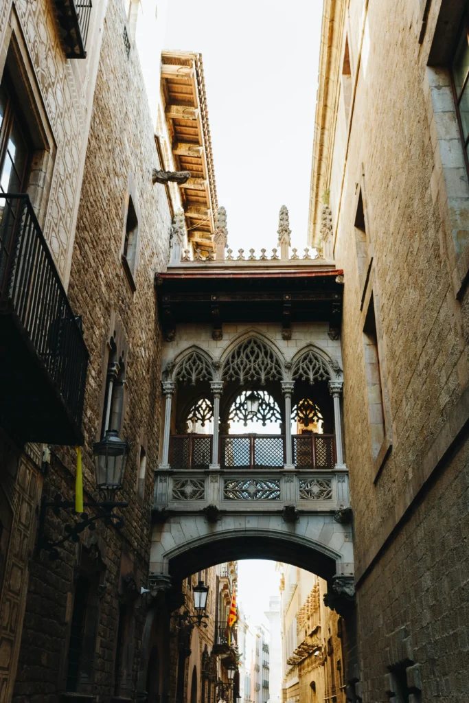 Un puente peatonal de piedra con arcos góticos conecta dos edificios históricos sobre una calle estrecha cerca de L'Auditori de Barcelona, en una ciudad europea.