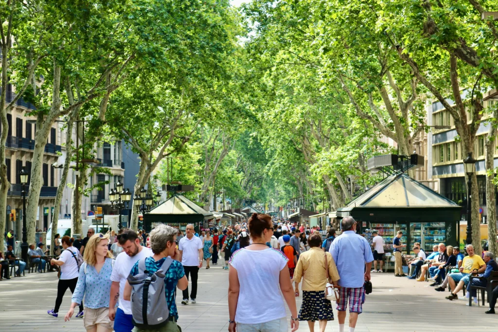 Una concurrida calle peatonal arbolada de Barcelona con gente paseando, pequeños quioscos y edificios a ambos lados bajo un dosel de hojas verdes, a pocos pasos de la renombrada sala de conciertos L'Auditori de Barcelona.