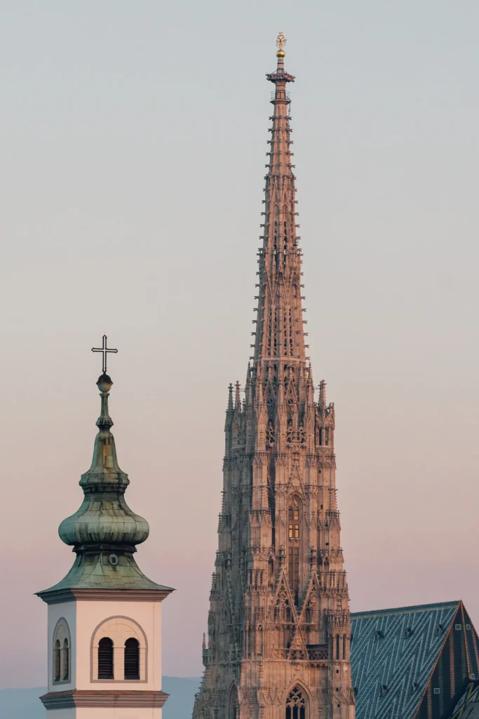 A church steeple with a cross and the tall, ornate spire of St. Stephens Cathedral in Vienna rise against a pale sky at sunset.