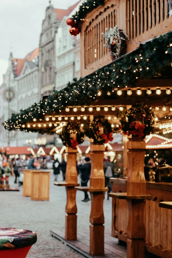 Dřevěné venkovní stánky na Václavském náměstí ozdobené girlandami a věnci, osvětlené světelnými řetězy, v pozadí procházející se lidé na dlážděné ulici v Praze.