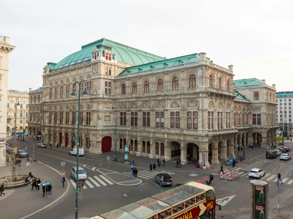 A large historic building, St. Stephens Cathedral, with a green roof and ornate stone facade sits at a busy city intersection with cars, pedestrians, and a tour bus nearby.