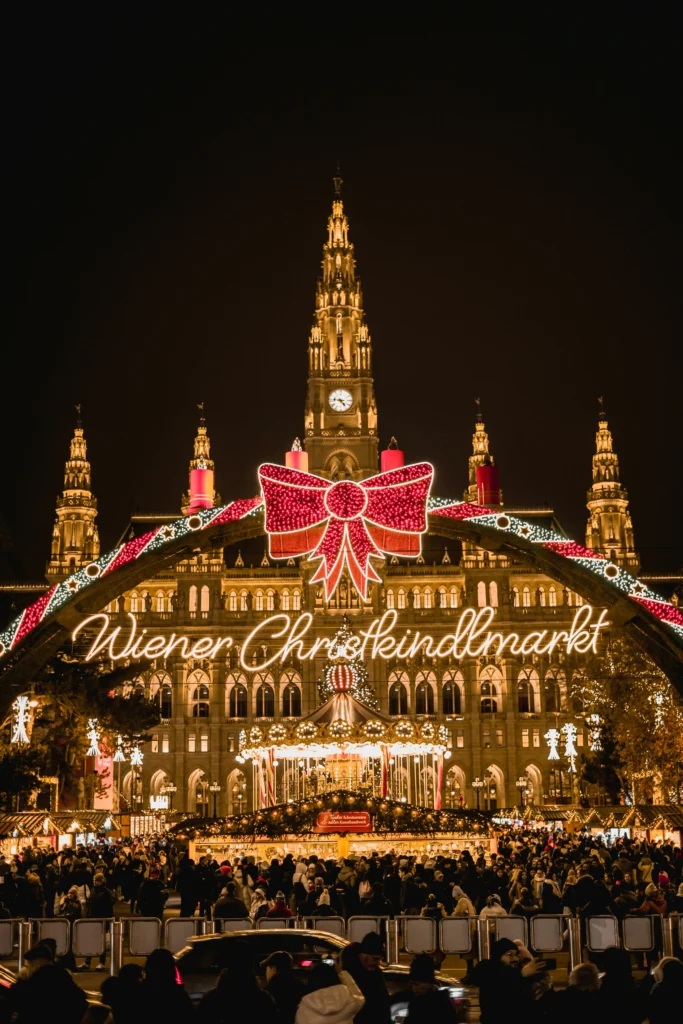 Das Wiener Rathaus ist in der Nacht mit festlichen Lichtern und einer großen roten Schleife für den Wiener Christkindlmarkt beleuchtet; Menschenmengen versammeln sich davor, nur einen kurzen Spaziergang vom ikonischen Stephansdom entfernt.