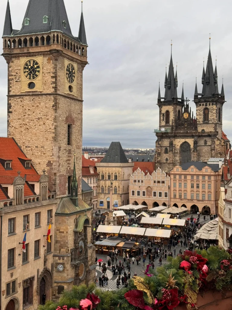 Pohled na Staroměstské náměstí s orlojem, historickými budovami, vánočním trhem a věžemi Týnského chrámu pod zataženou oblohou. V popředí u Václavského náměstí je vidět sváteční výzdoba.