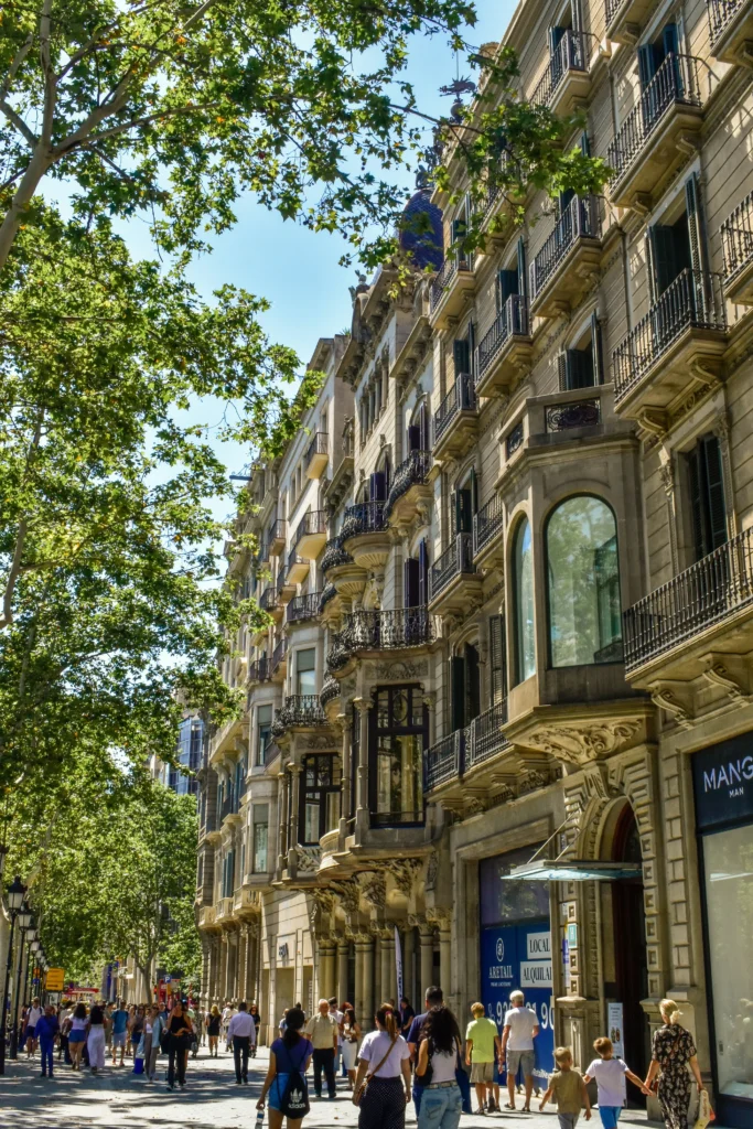 People stroll along a tree-lined street in Barcelona next to historic multi-story buildings with balconies and shops at ground level, enjoying the sunny day.