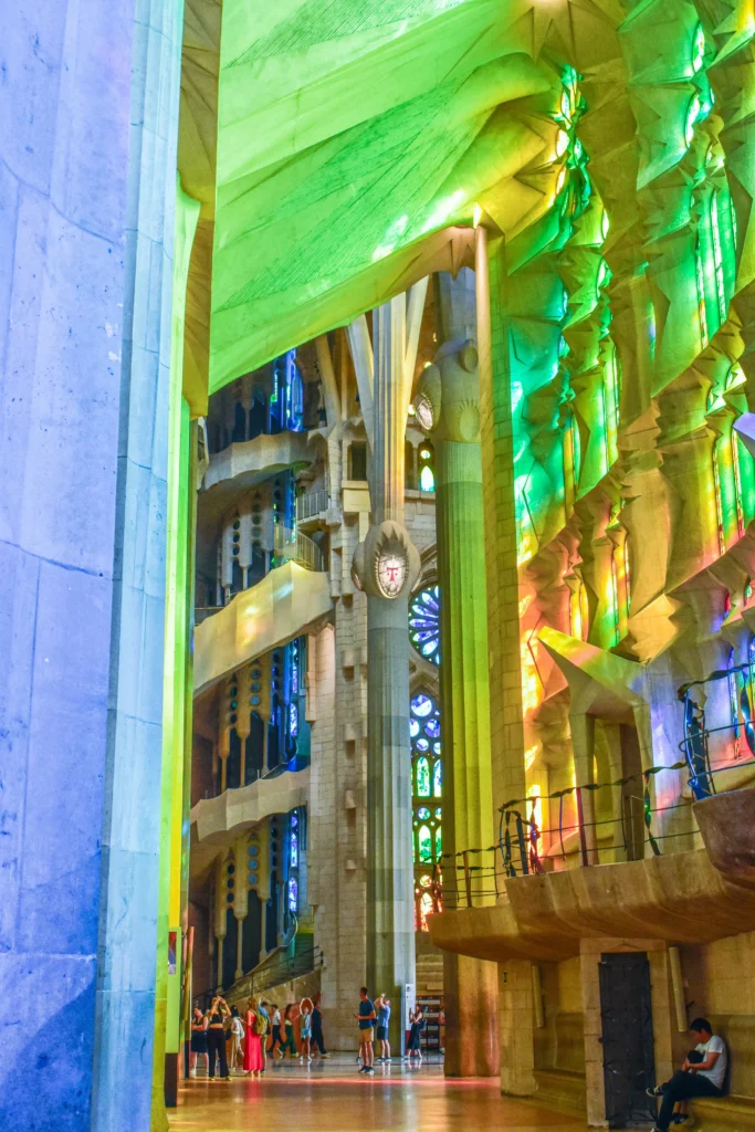 Vista interior de la Sagrada Familia con vidrieras de colores que proyectan luz verde y amarilla, altas columnas y visitantes de pie junto a la pared, un ambiente que recuerda al de las grandes salas de conciertos como L'Auditori de Barcelona.