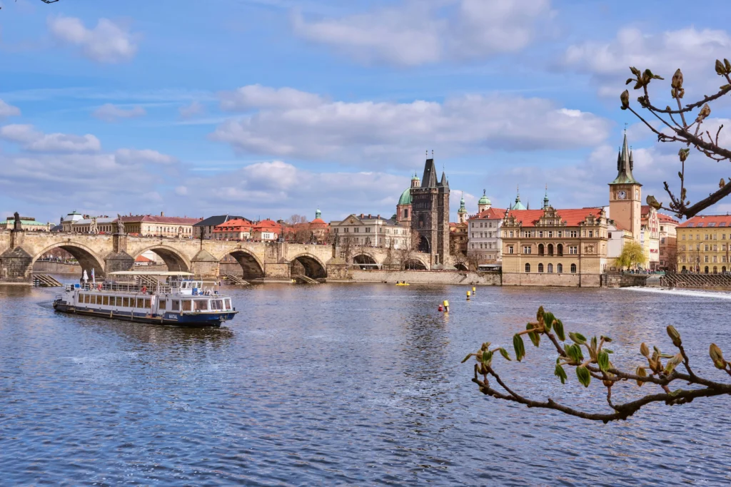 Loď pluje po řece před historickým kamenným mostem a starými městskými budovami nedaleko Václavského náměstí, pod částečně zataženou oblohou.