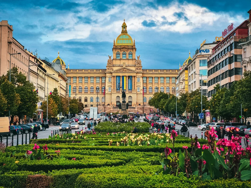 A large ornate building with a green dome stands at the end of Prague’s bustling Wenceslas Square, its boulevard lined with cars, trees, and flower beds under a cloudy sky.