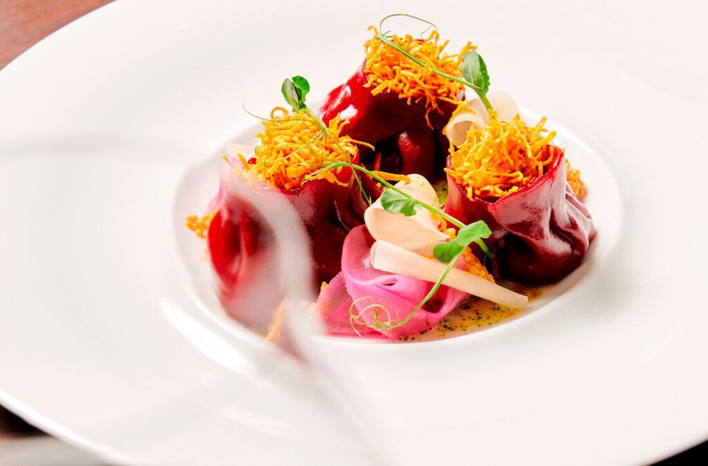 A white plate with a gourmet dish featuring beetroot ravioli, thin vegetable slices, microgreens, and crispy golden shreds—perfect for impressing guests at events or your next important meeting.