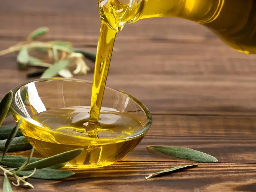 Olive oil being poured from a glass bottle into a small glass bowl, with olive branches on a wooden surface—an inviting scene that highlights how to make olive oil from scratch.