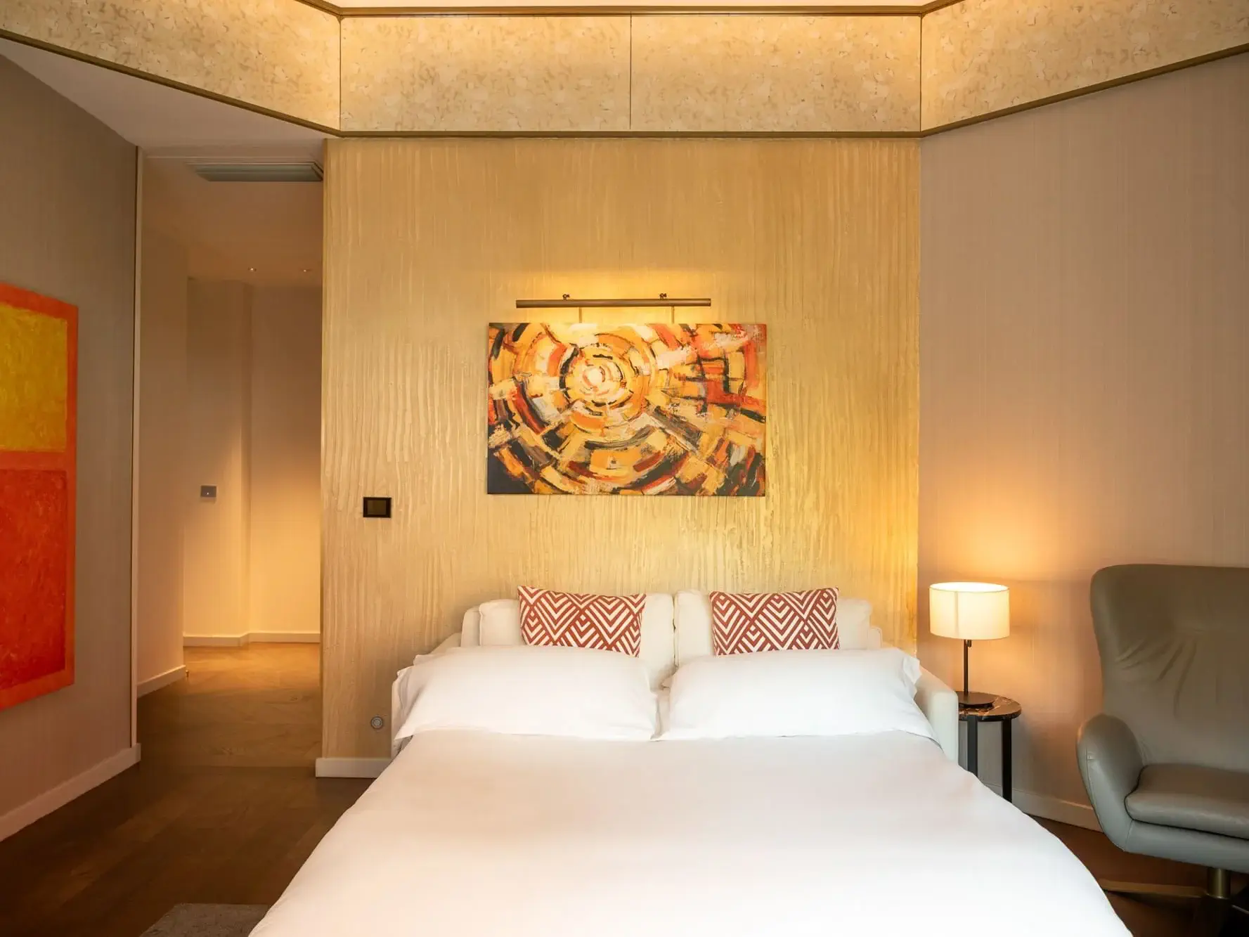 Un llit ben fet amb roba de llit blanca i coixins amb estampats vermells es troba contra una paret daurada amb obres d'art abstractes a sobre, oferint un toc acollidor a aquesta suite familiar, flanquejada per una làmpada i una cadira moderna de color beix.