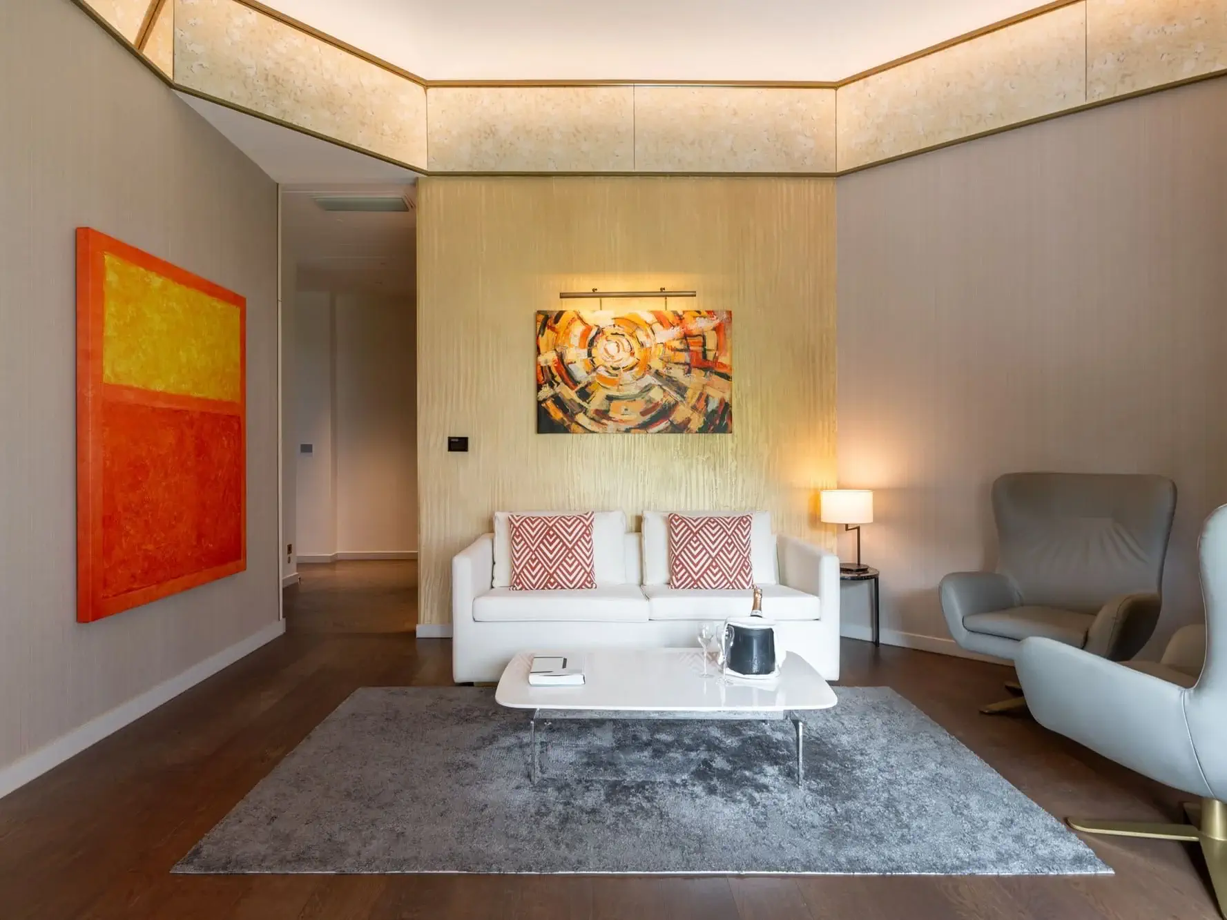 Sala d'estar moderna a la suite familiar Almanac amb un sofà blanc, art abstracte a la paret, catifa grisa, taula de centre i butaca grisa. Una il·luminació càlida i unes parets neutres creen una atmosfera contemporània.