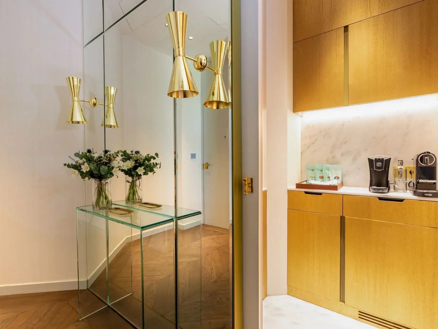 Un passadís modern de la suite familiar Almanac compta amb una consola de vidre, dos gerros de flors, aplics de paret daurats i una cuineta amb armaris daurats, cafeteres i un revestiment de marbre.