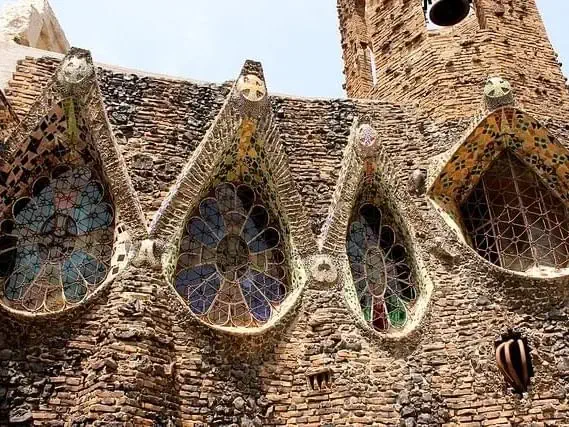 Close-up of a rustic brick building facade featuring three large, pointed stained-glass windows with colorful designs and intricate mosaic details above each window, showcasing the vibrant influence of Catalan Modernism.