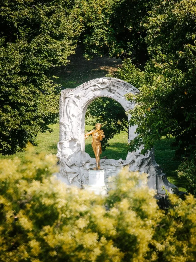 A golden statue of Strauss stands under a white arch adorned with relief figures, surrounded by lush green trees—a Romantic Tribute amid nature.