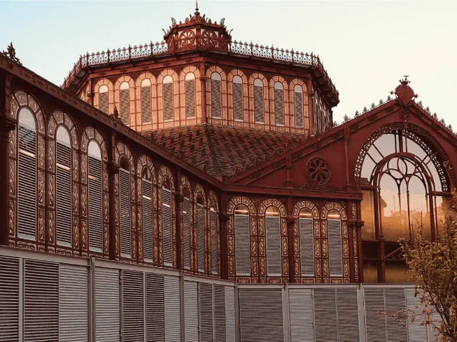 A historic Barcelona building with intricate ironwork and arched windows reflects warm light; the lower section features modern metal panels, inviting you to savour the earth’s unique blend of old and new.